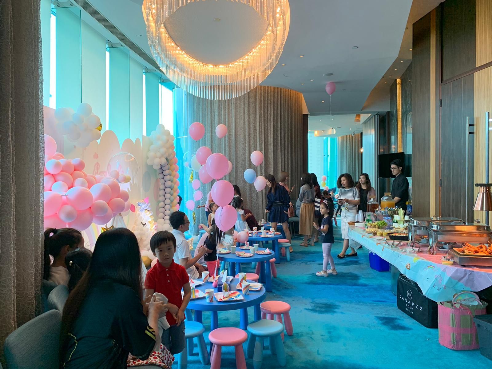 A party with pink and blue balloons, children at small tables, and a buffet in a room with a large window.