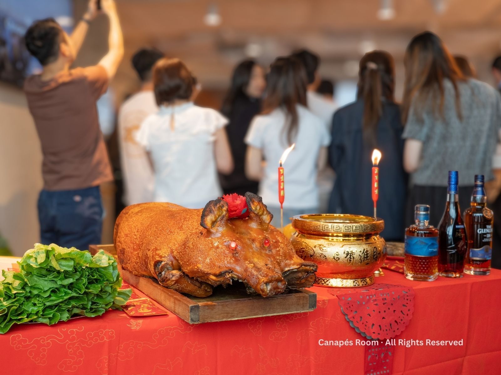 Roasted pig on table with candles, liquor, and onlookers in a brightly lit room.