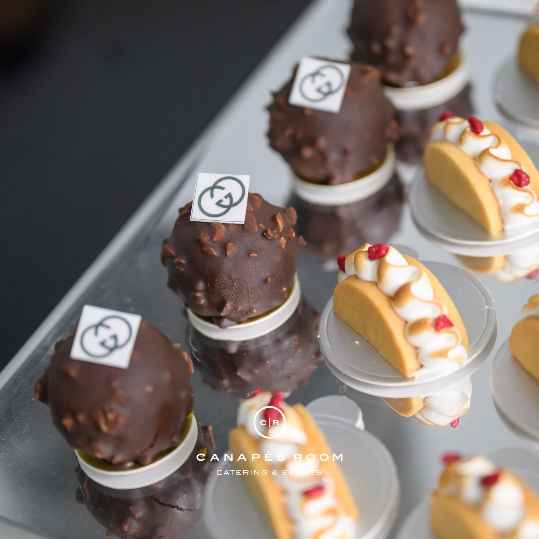Mini desserts on a mirrored tray: chocolate spheres with Gucci logo, and miniature tacos with sauce and berries.
