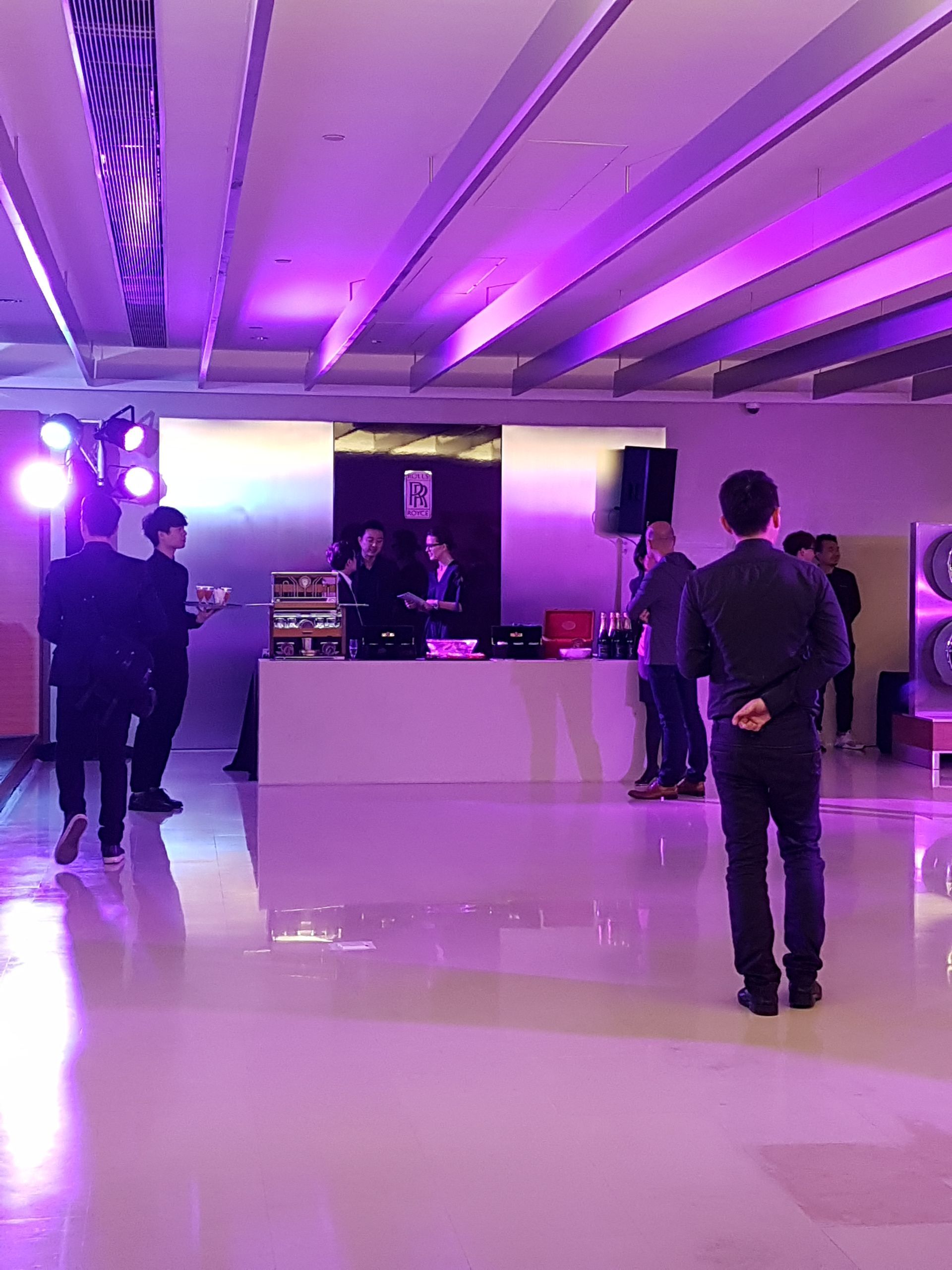 People in a room with purple lighting. Some are standing by a table, others are watching.
