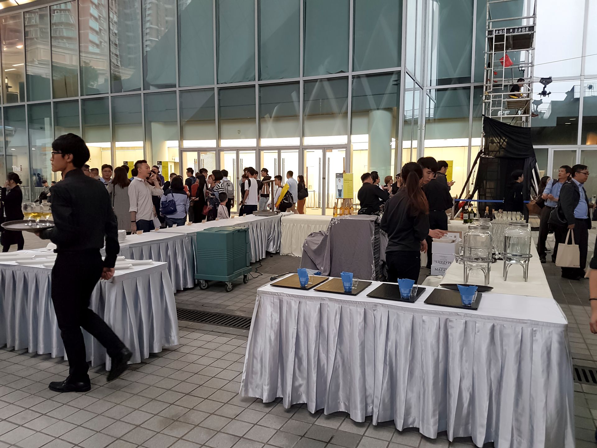 People at an event with food and drinks served on covered tables, servers in black, and a glass-walled building.