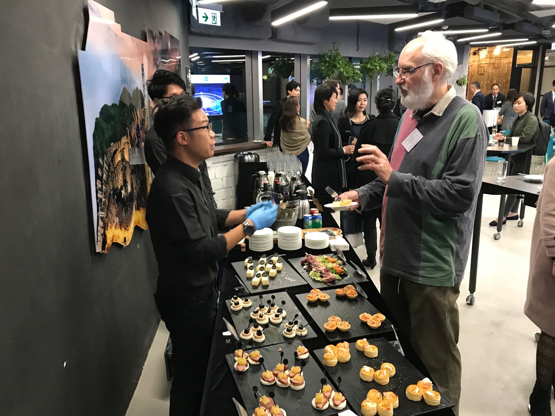 A server in black uniform serves appetizers to a man with a beard at an event with other attendees.