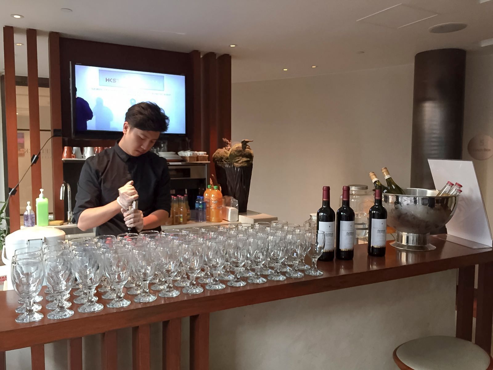 Bartender preparing drinks behind a bar with many glasses; bottles of wine and champagne visible.