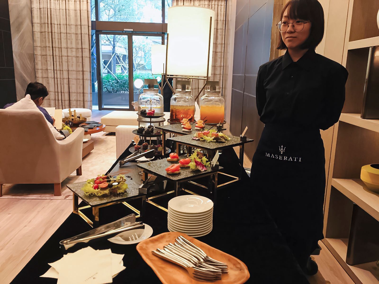 Woman in black apron stands at a buffet table with drinks, snacks, and serving utensils.