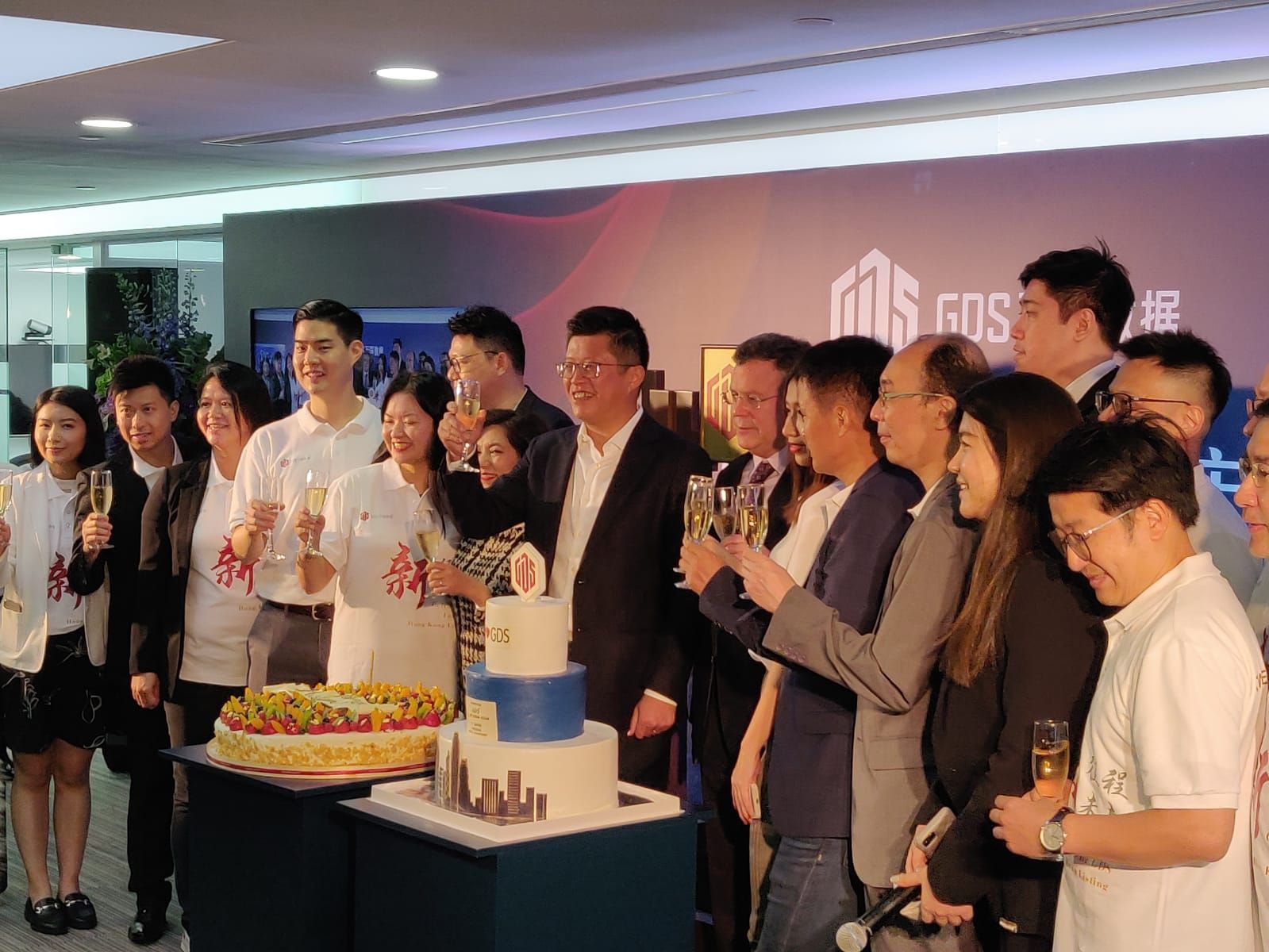 Group celebrates with cake and champagne; party indoors with company logo in background.