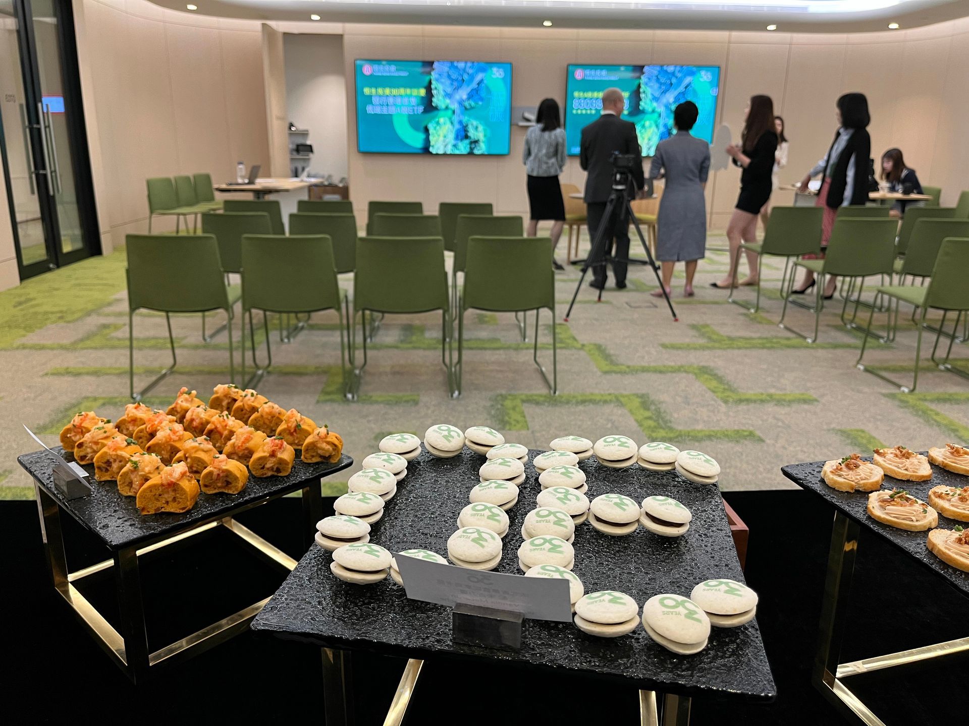 Reception room with snacks, chairs, and people in front of screens.