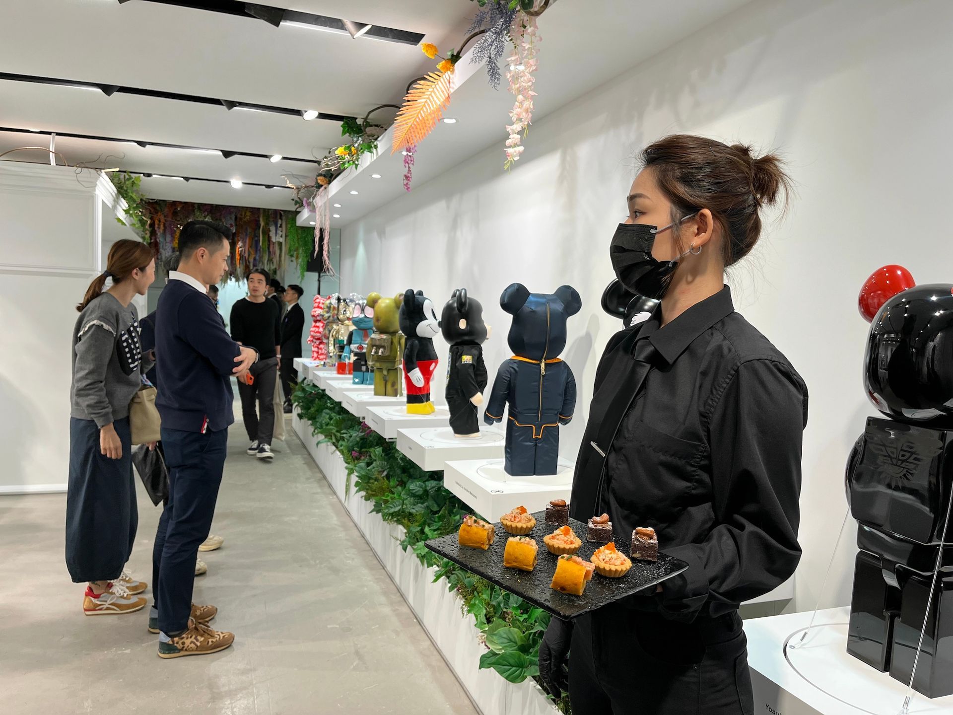 People view toy figures in a gallery; a server offers food.