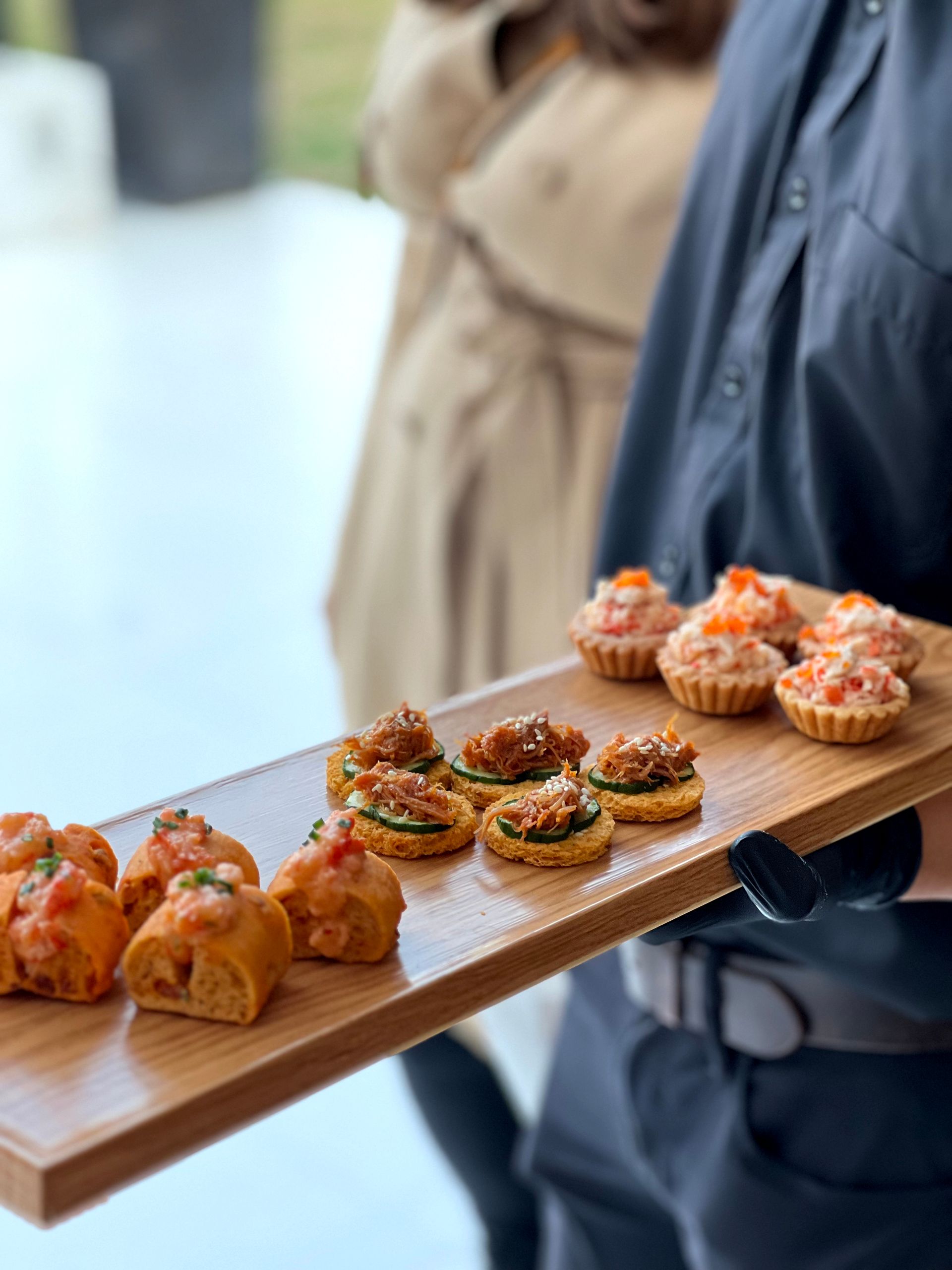 A server holding a wooden tray with various appetizers. The background is blurred with people.