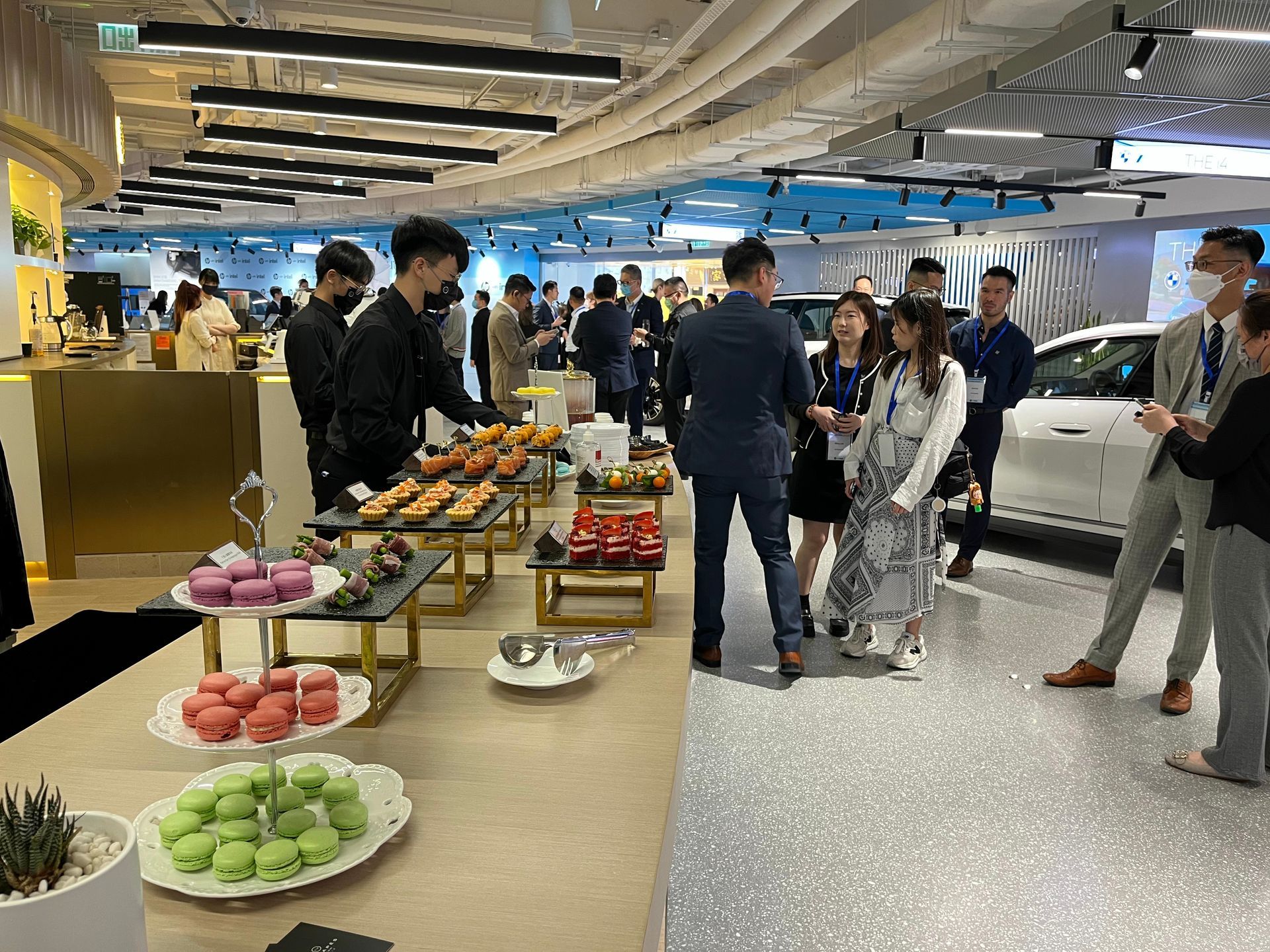People at a reception with food and a car display in a bright, modern venue.