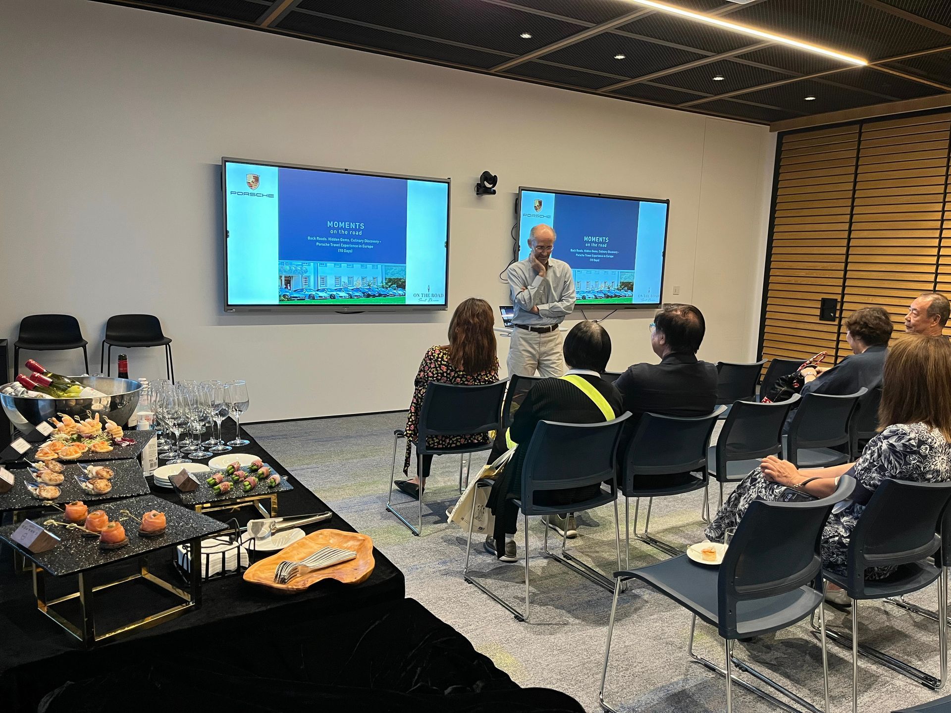 A man gives a presentation to a seated audience, with two screens displaying information. A food table is in the foreground.