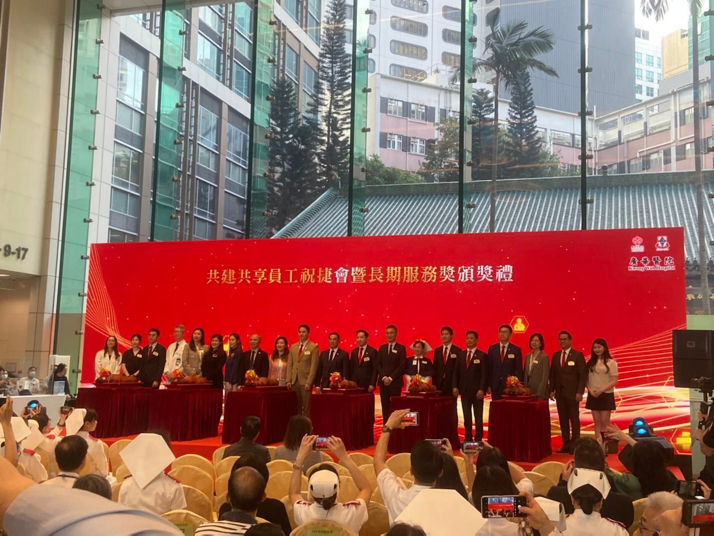 Group of people at an event standing behind tables and a large red banner.