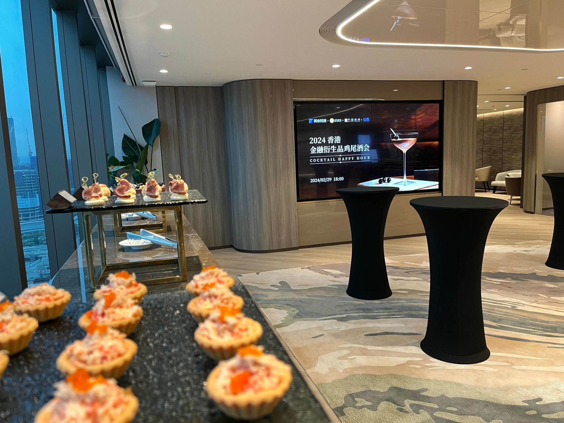 Buffet table with appetizers. Cocktail tables in a room with a large screen and floor-to-ceiling windows.