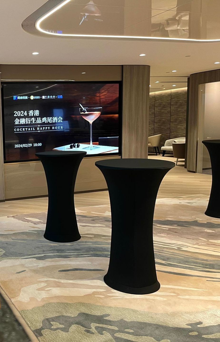 Black cocktail tables on patterned carpet, in front of a screen displaying a cocktail advertisement, interior setting.