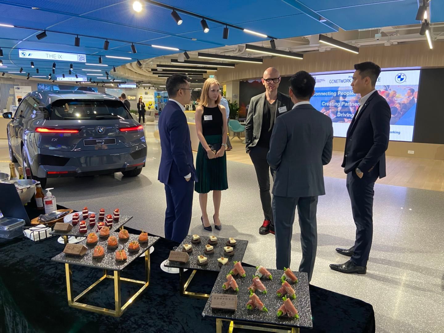 People in suits and dress at an event with a vehicle, dessert table, and blue and white decor.