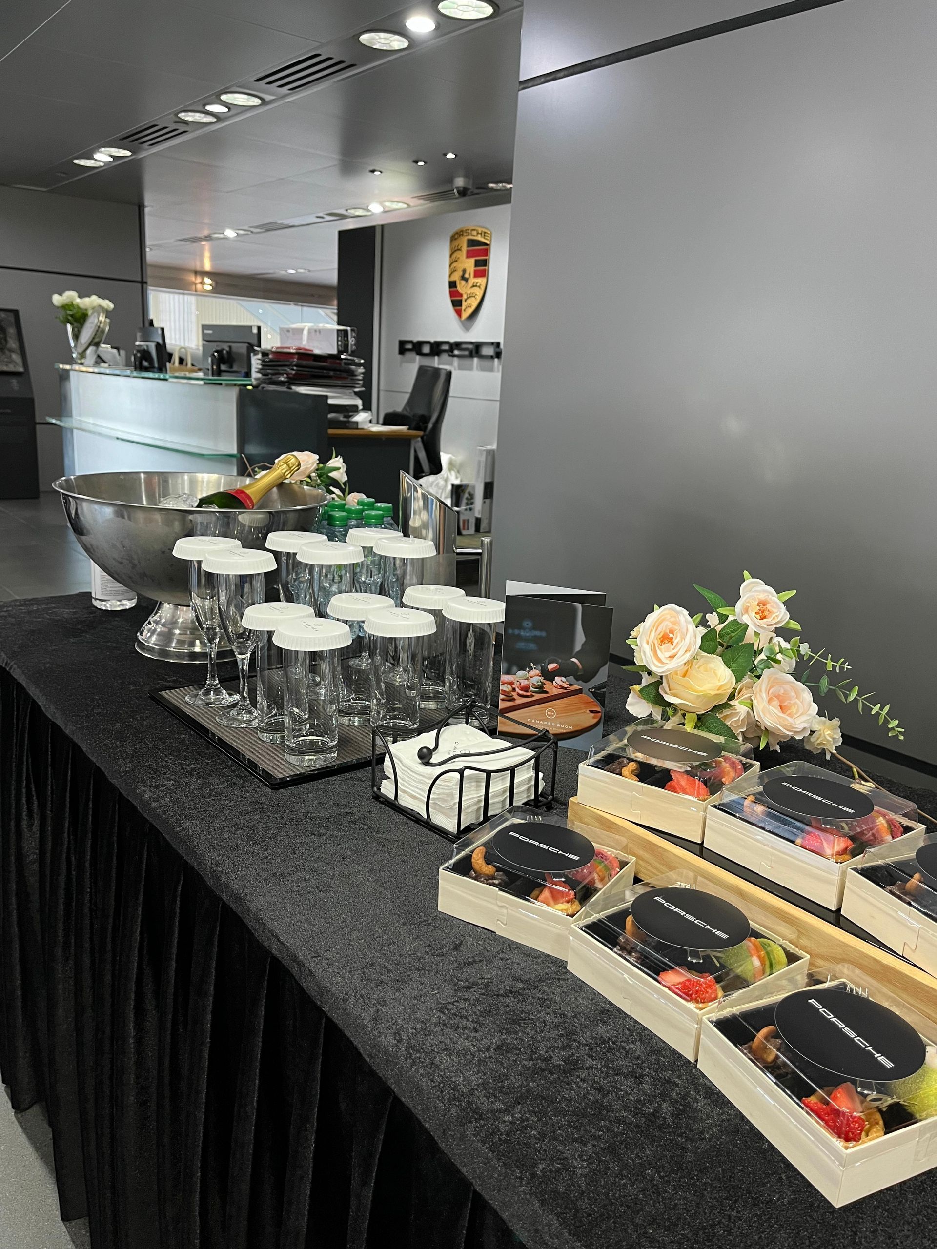 Buffet table with desserts, drinks, and flowers, set up in a Porsche dealership.