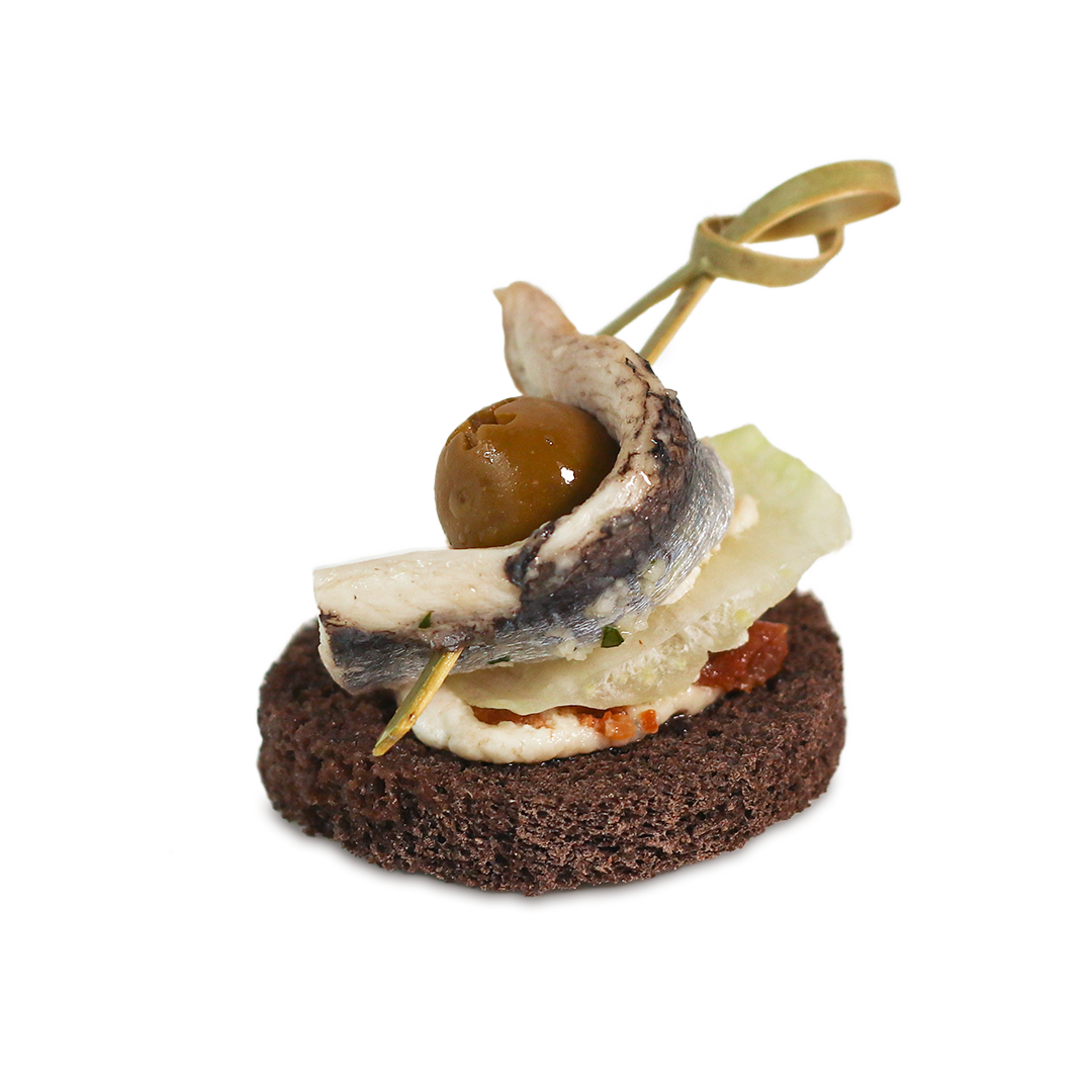 Appetizer with sardine, olive, and greens on a dark bread base, held together with a wooden skewer.