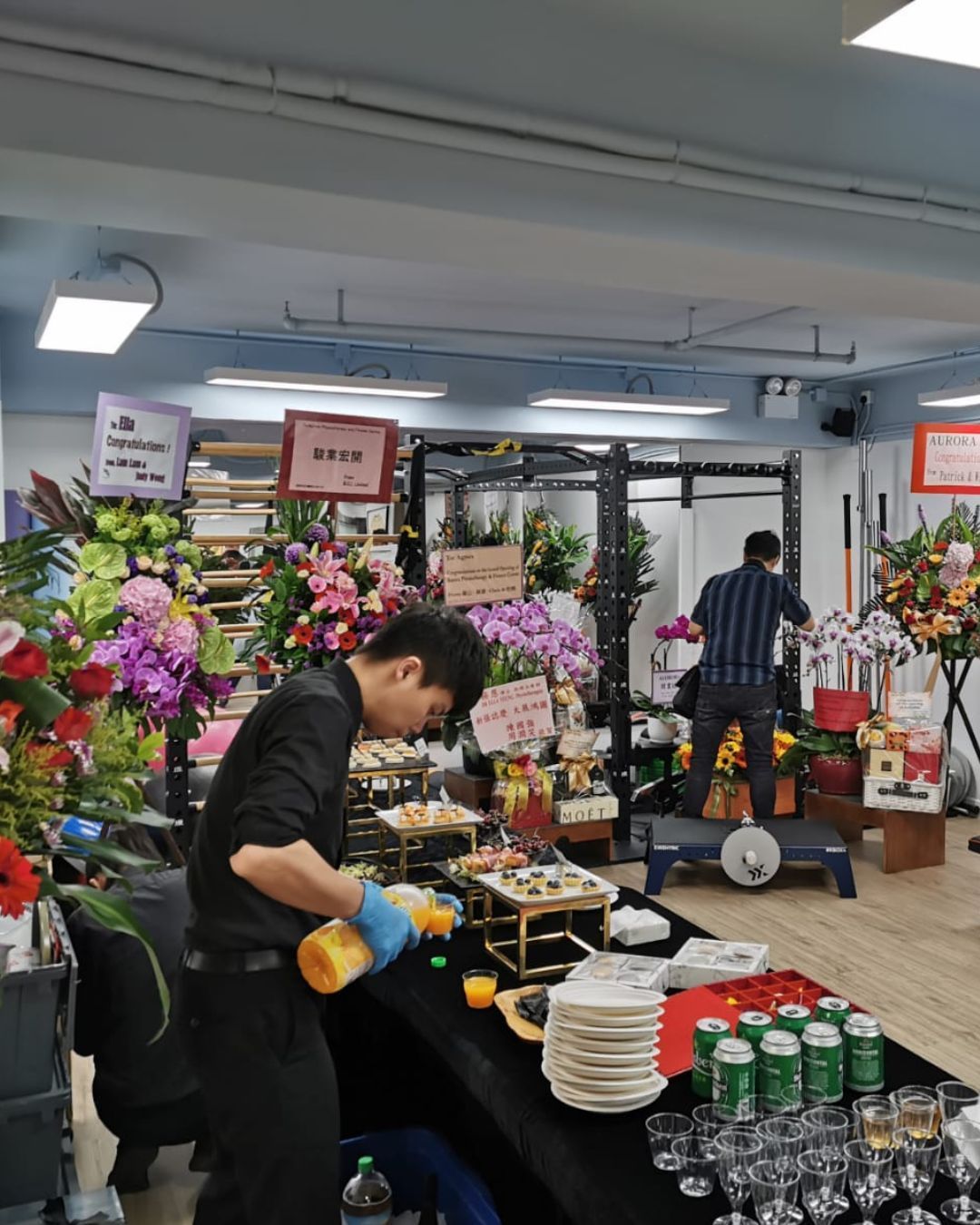 A man prepares food at a table adorned with drinks and snacks. Gym equipment and flower arrangements fill the background.
