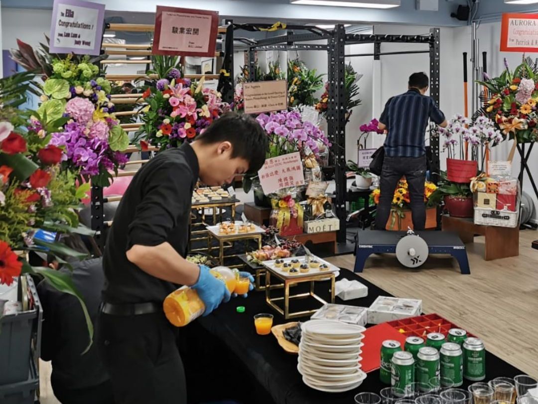 Two men prepare refreshments at an event. A table laden with food and drinks sits near floral arrangements and gym equipment.