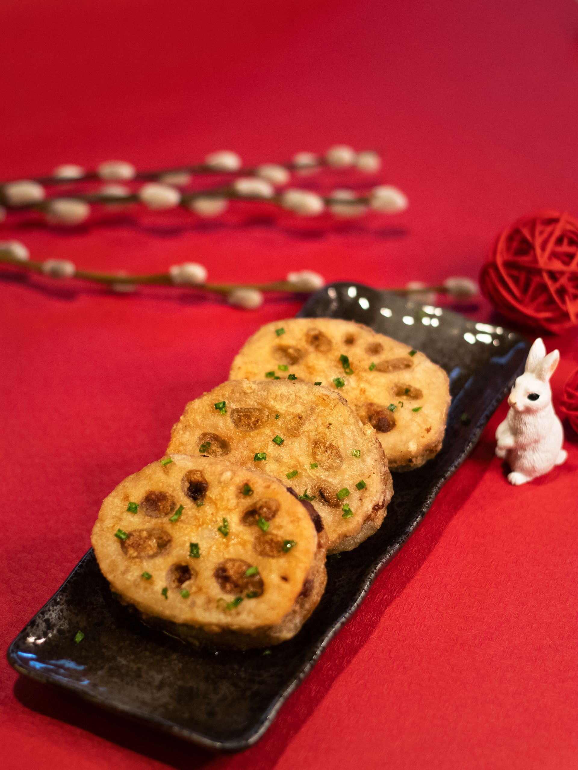 Fried lotus root slices on a black plate, red background, white bunny figurine, and willow branches.