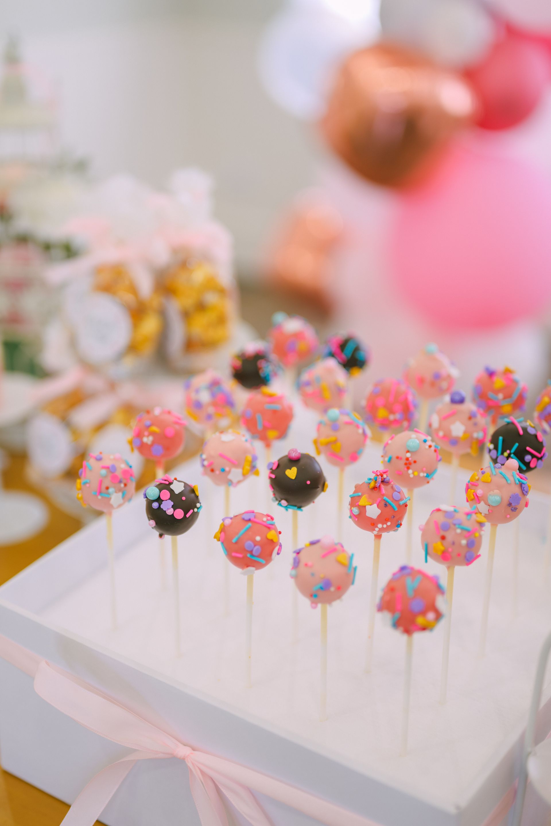 Cake pops on sticks in a white box, decorated with sprinkles. Pink and gold balloons in the background.