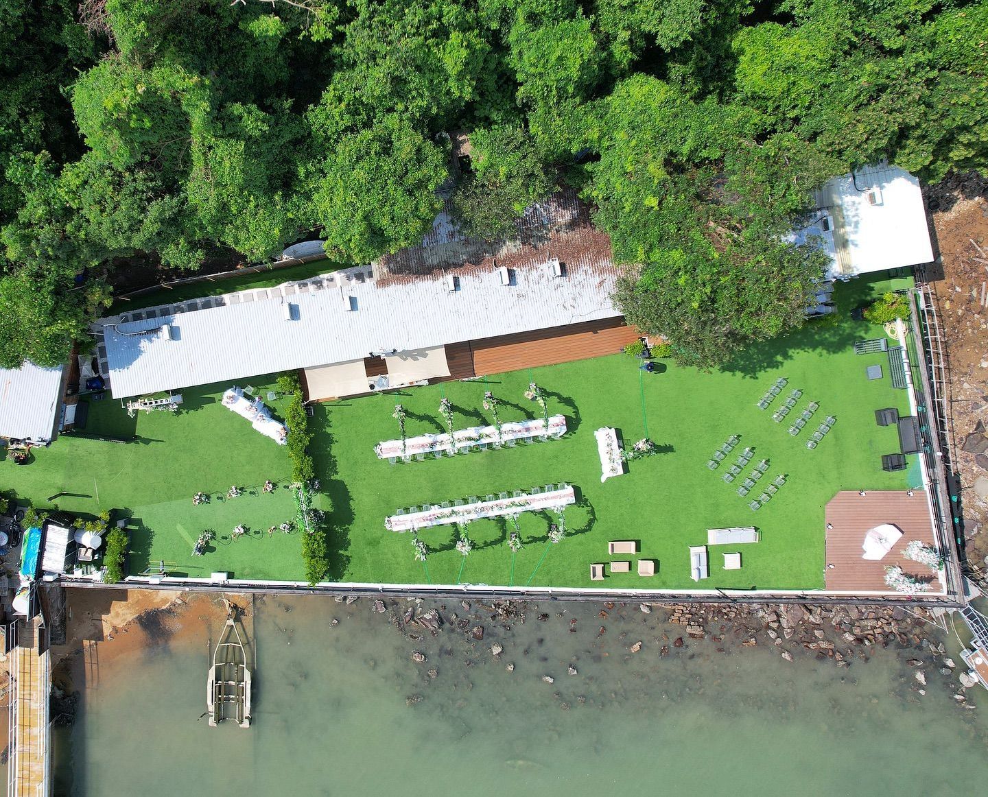 俯瞰圖：活動場地位於湖岸邊，綠草茵茵，樹木蔥鬱，建築物林立，水面波光粼粼。白色桌椅錯落有致地擺放著。