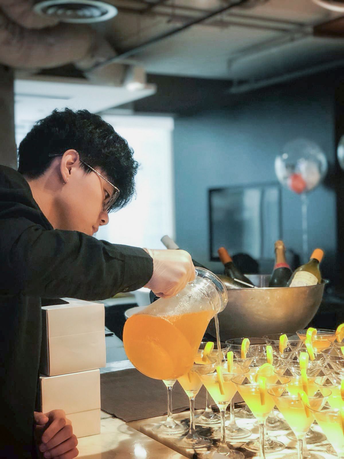 Person pouring orange liquid into martini glasses at a bar.