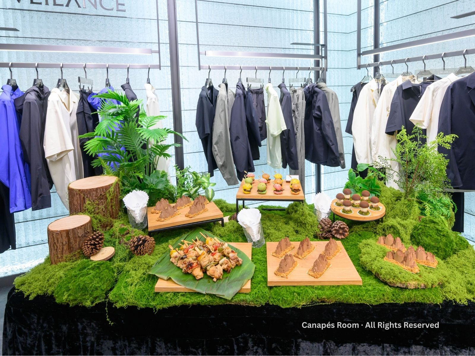 Food display on moss-covered table with clothing rack background. Various snacks on wooden boards and leaves.