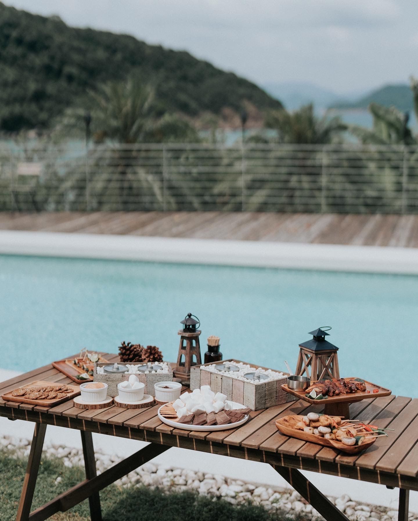 Wooden table with snacks and lanterns by a pool with a mountain backdrop.
