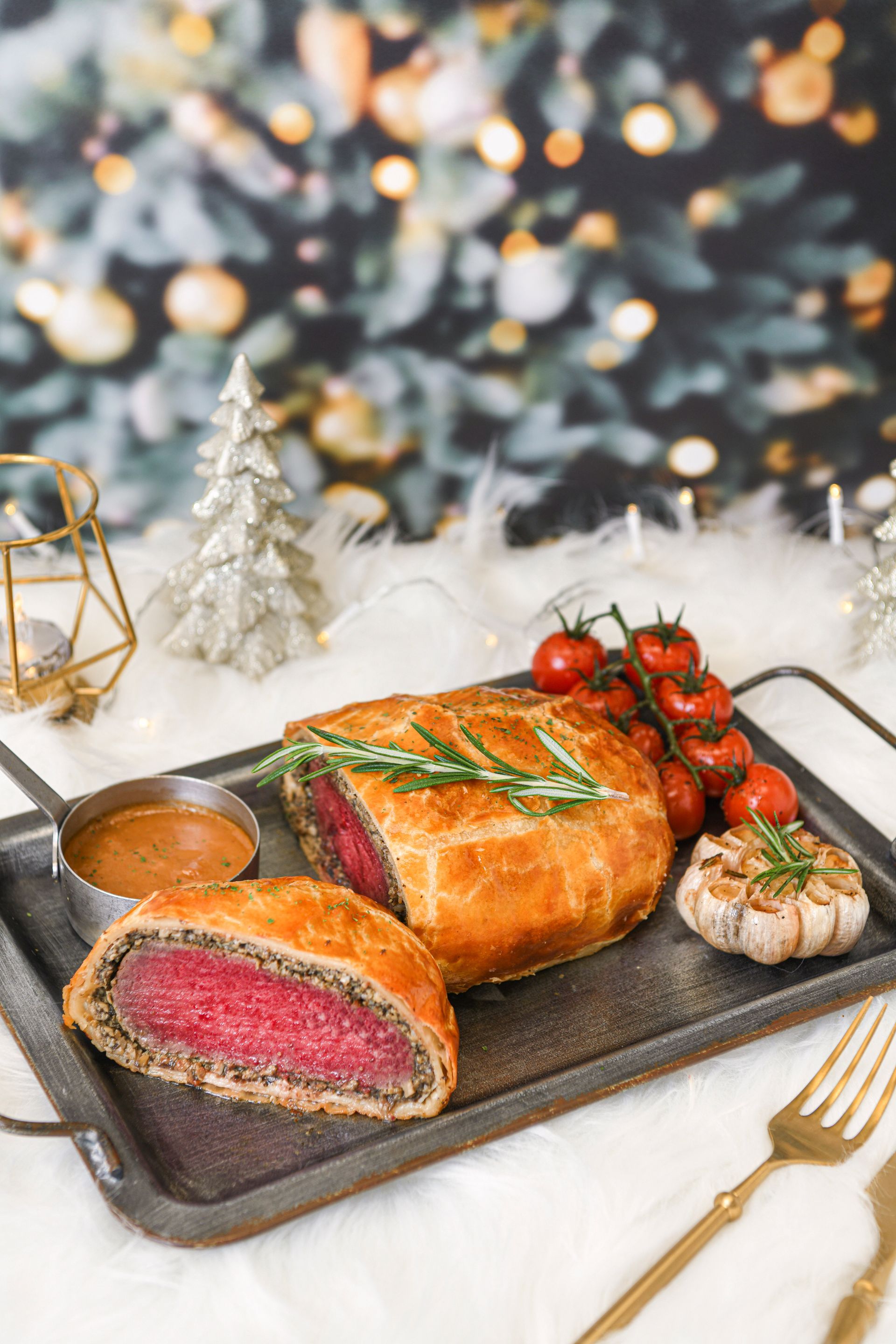 Beef Wellington with tomatoes, garlic, and sauce on a tray, Christmas lights in the background.