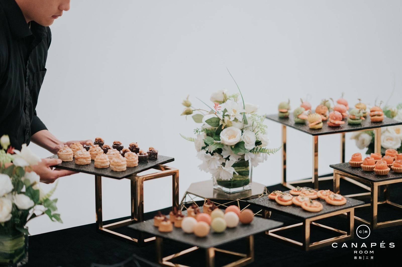 Person setting out appetizers on black and gold serving stands, with floral centerpiece on white background.