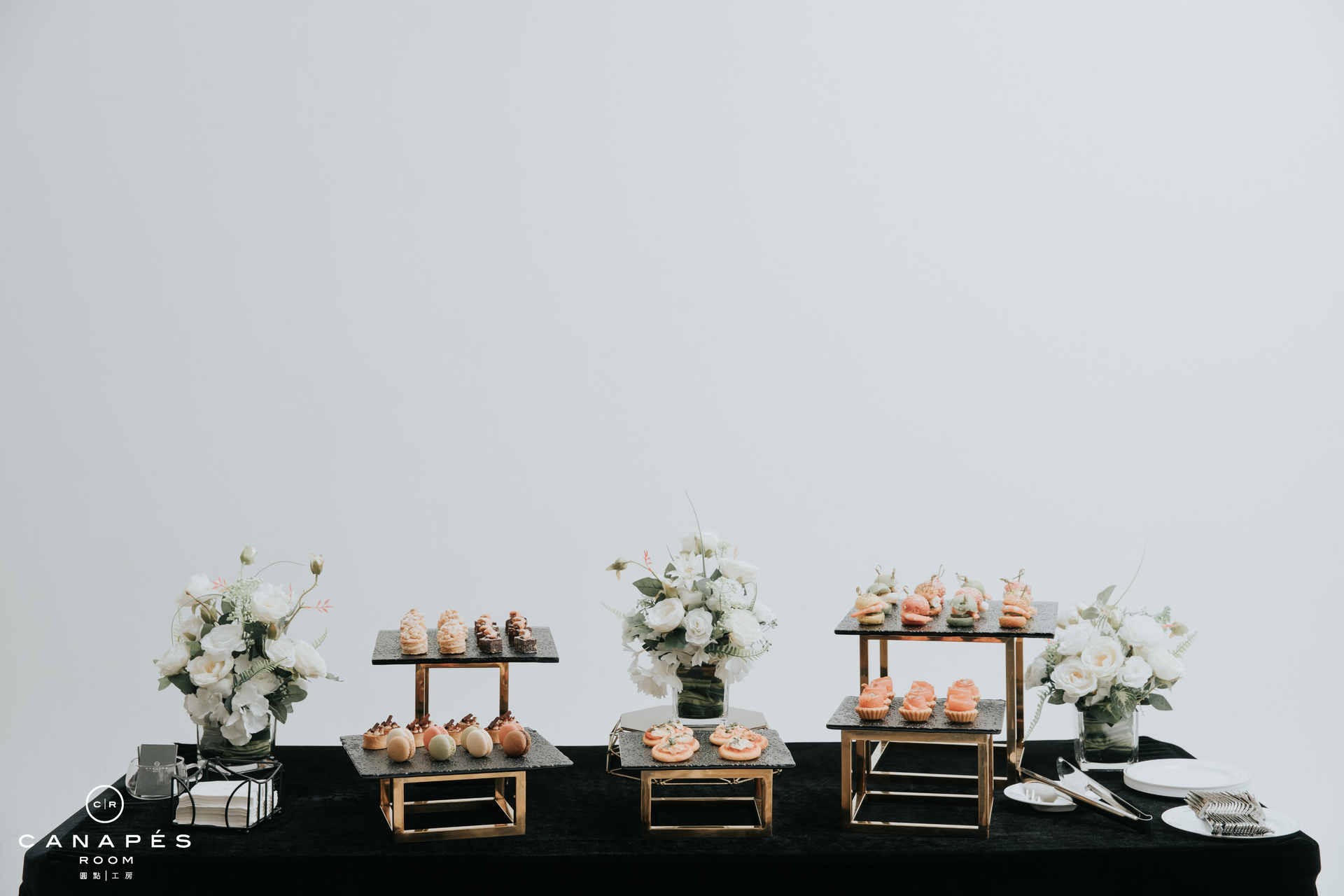 Dessert table with treats and white floral arrangements on a black tablecloth against a white background.