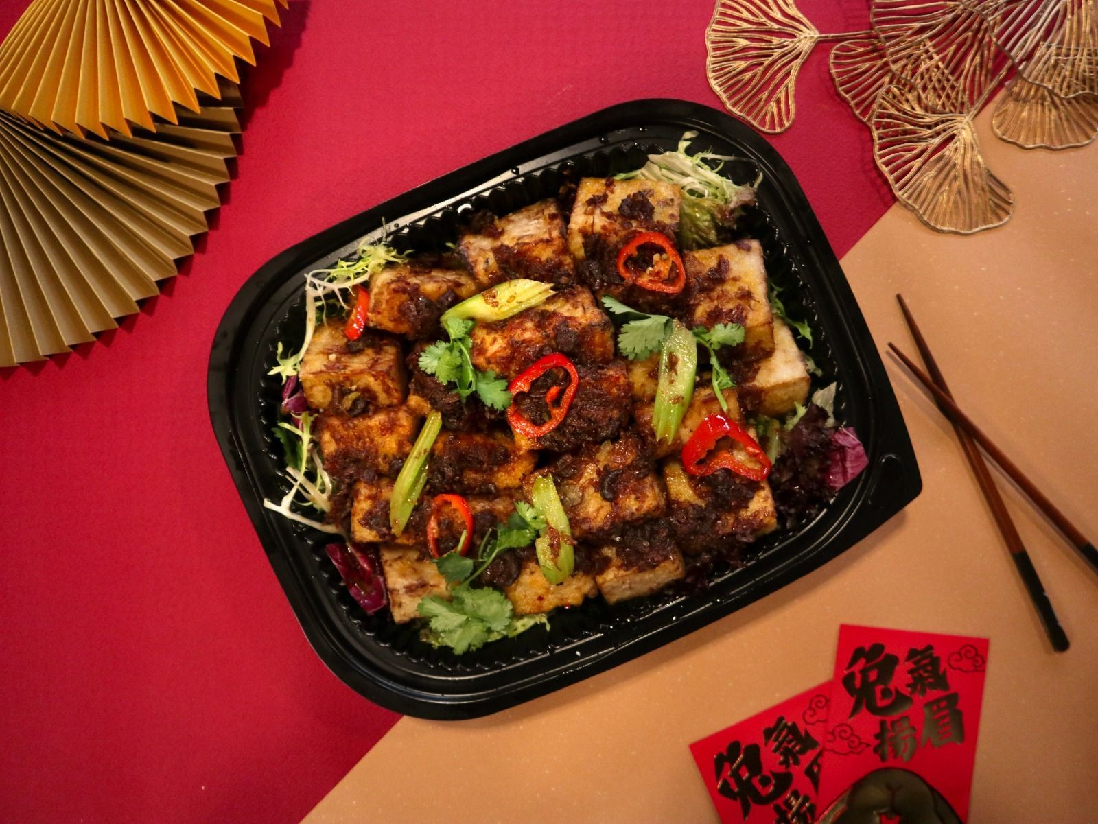 Tofu dish with chili peppers and herbs in a black container, chopsticks, and decorations on a red surface.