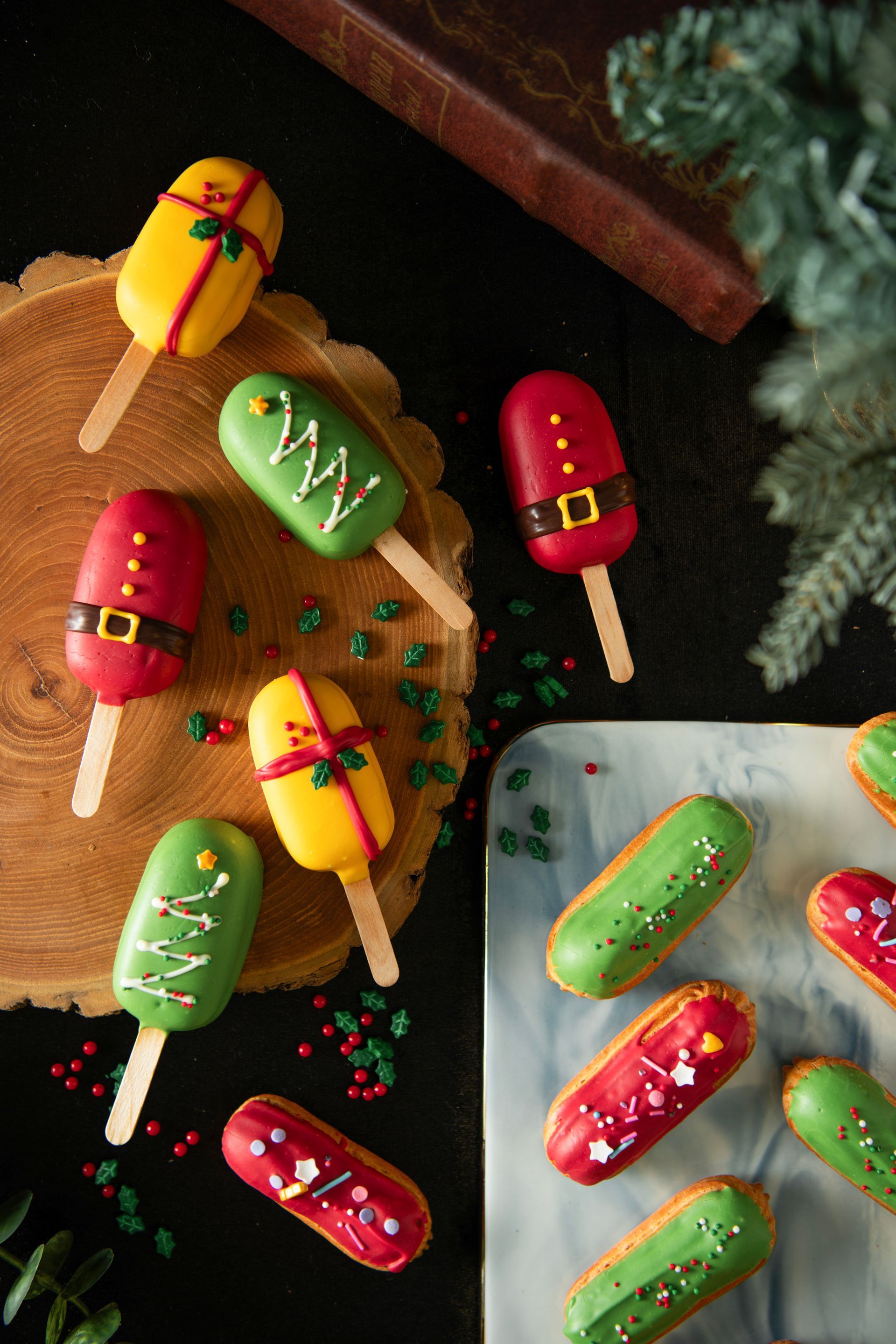 Christmas-themed desserts: Cake pops and eclairs decorated with colorful icing on wooden and marble surfaces.