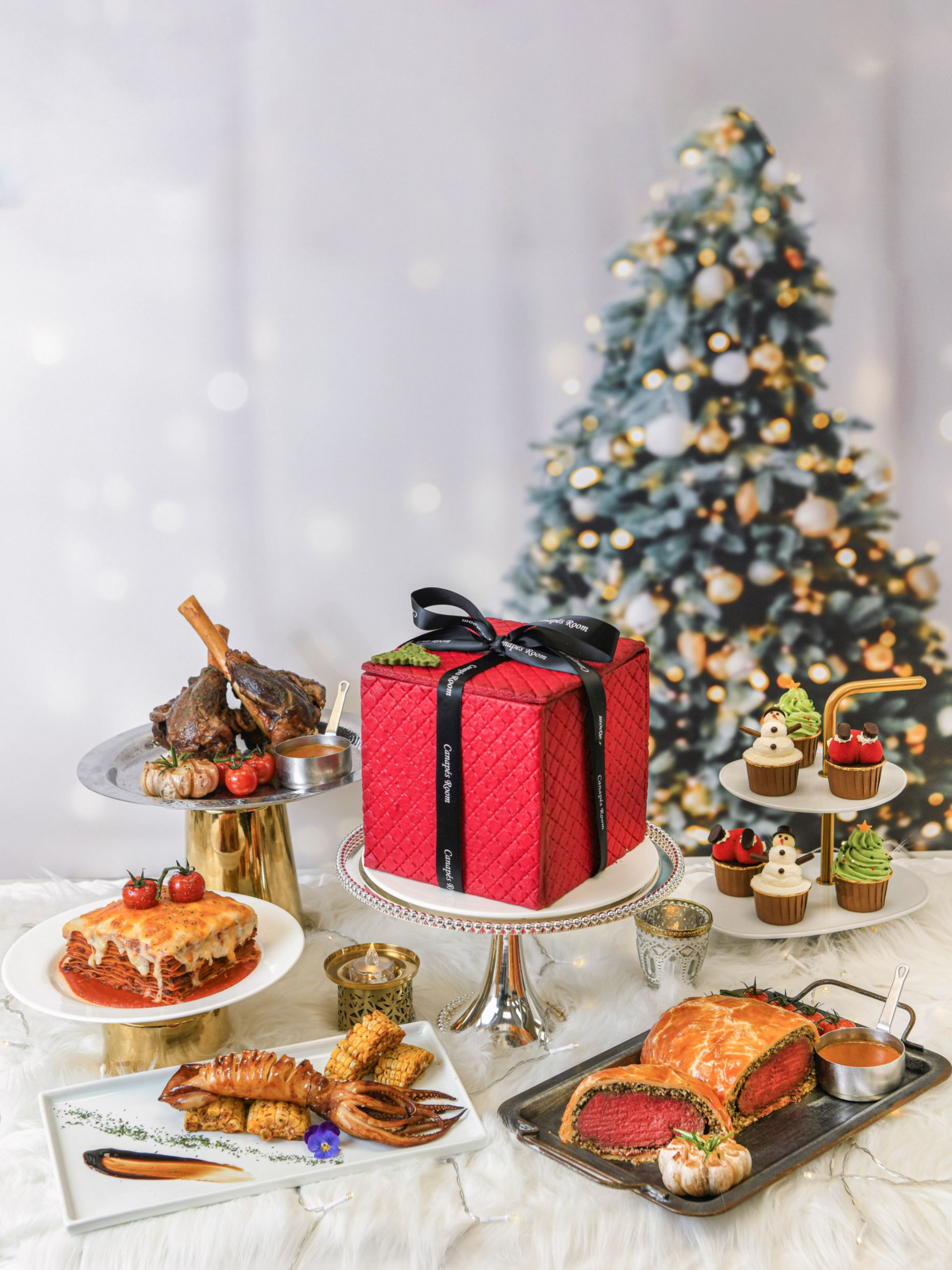 Christmas feast: lamb, lasagna, cake, Wellington, cupcakes, tree, and festive decor on a white table.