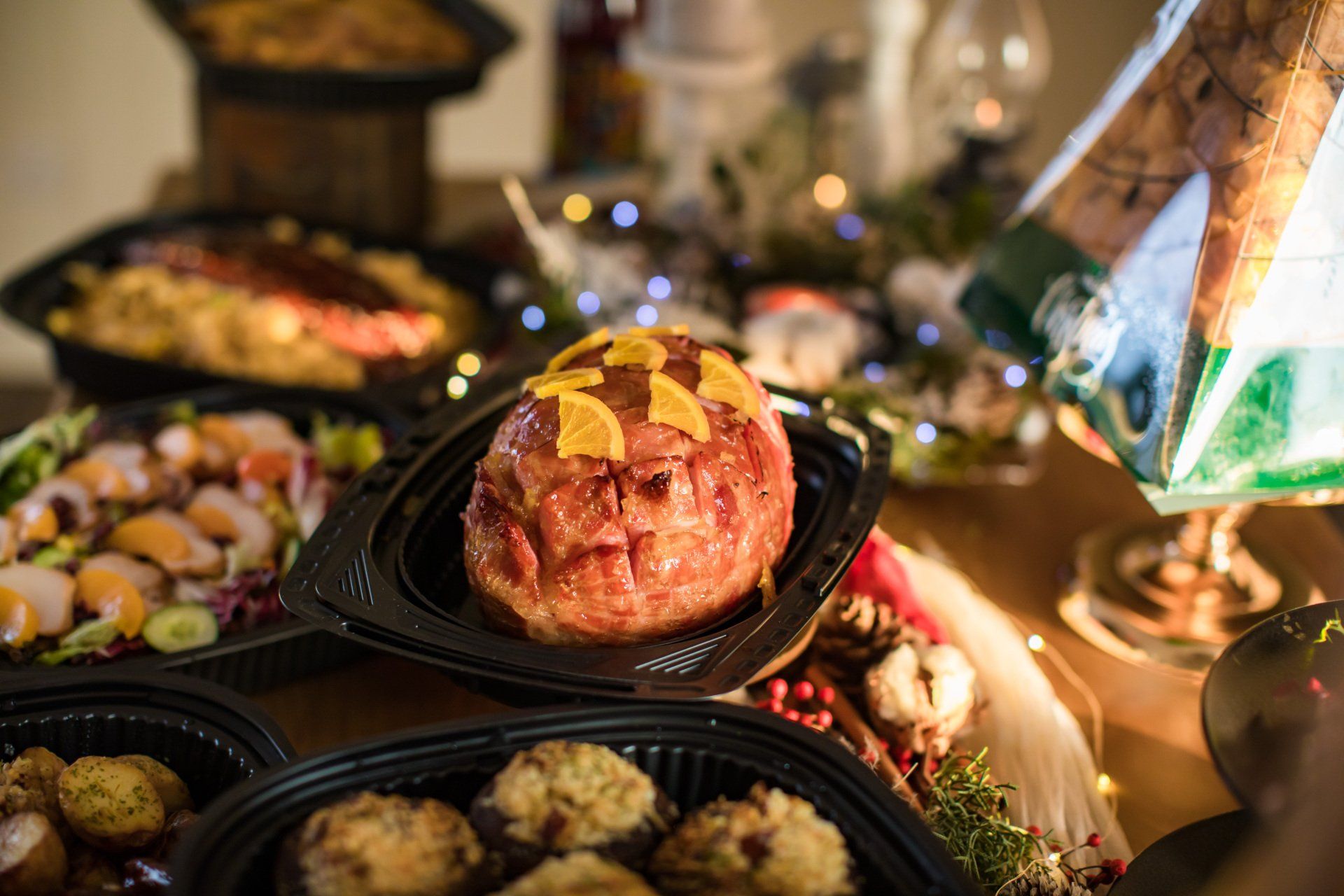 Holiday meal spread featuring ham with orange garnish, vegetables, and potatoes in black containers.