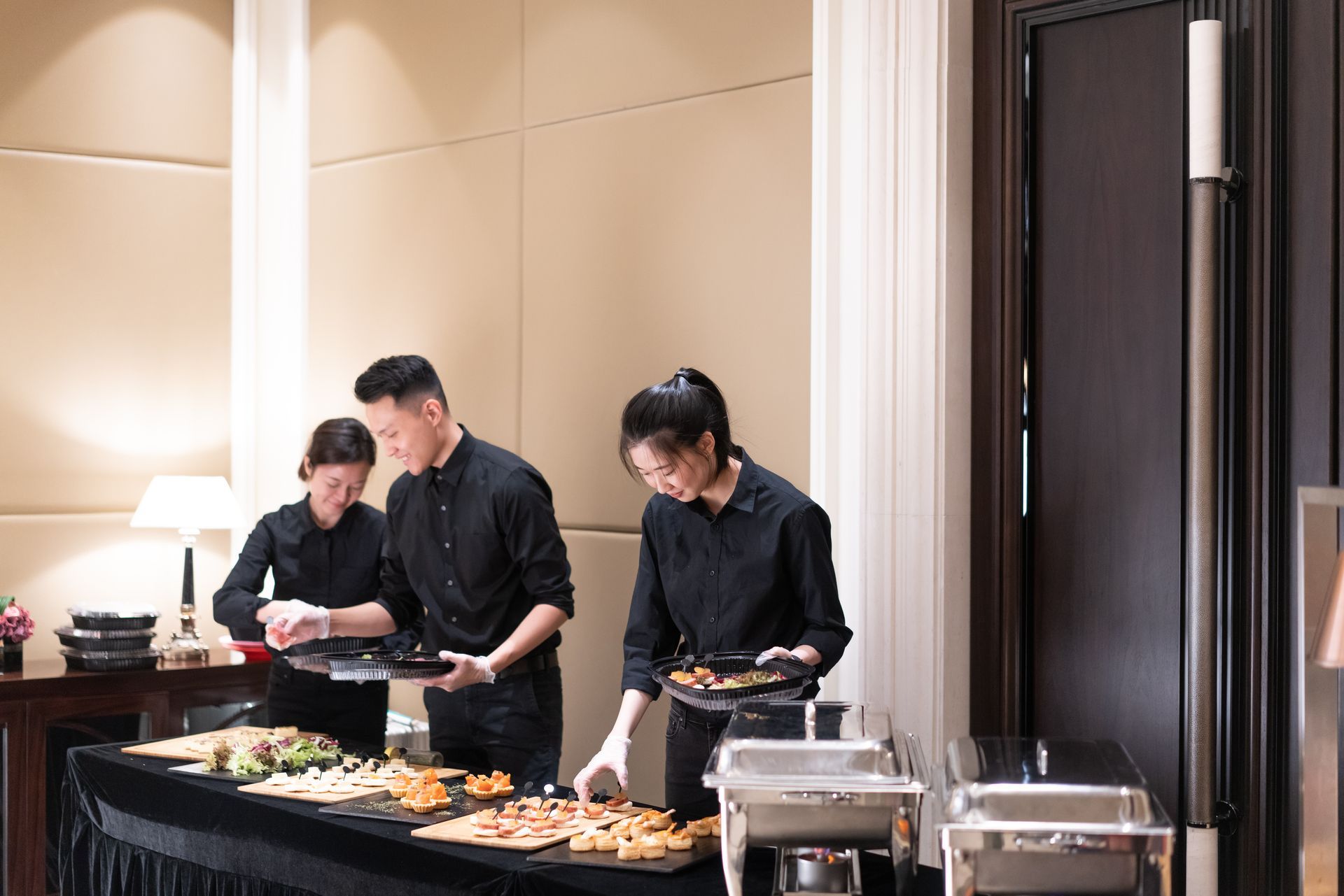 Three servers in black outfits arrange food on a buffet table in an event space.