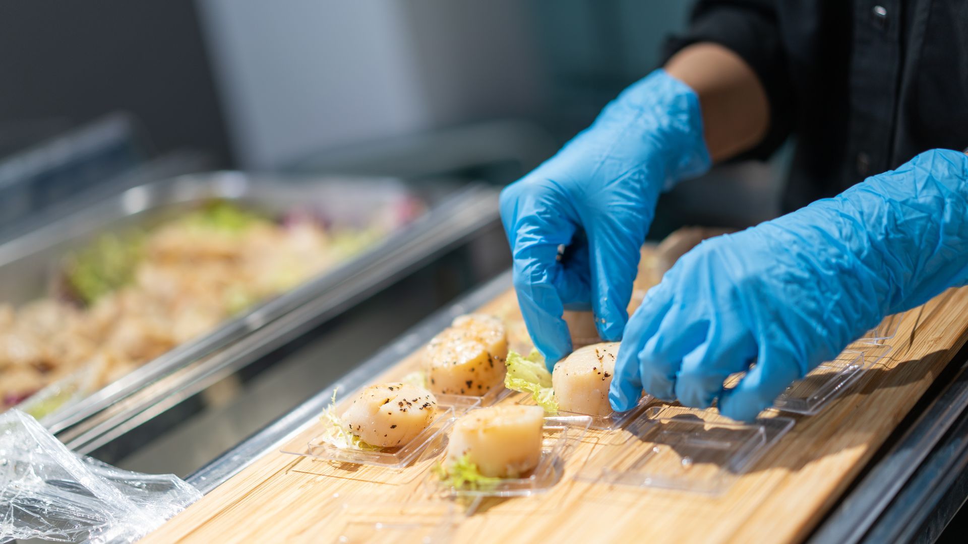 A person wearing blue gloves arranges seared scallops on small plates, next to a buffet tray.