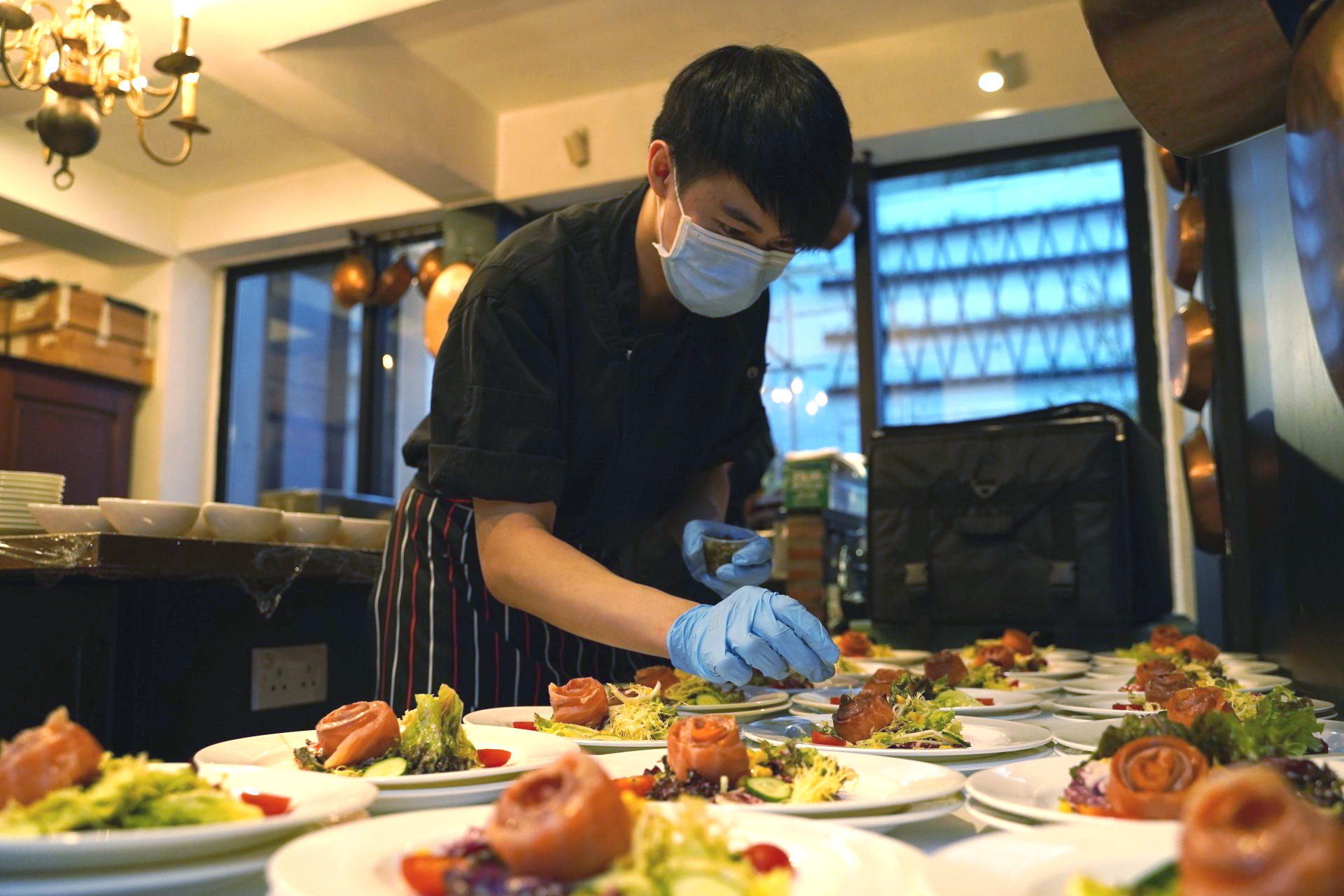 Chef wearing a mask and gloves prepares plates of food in a restaurant setting.