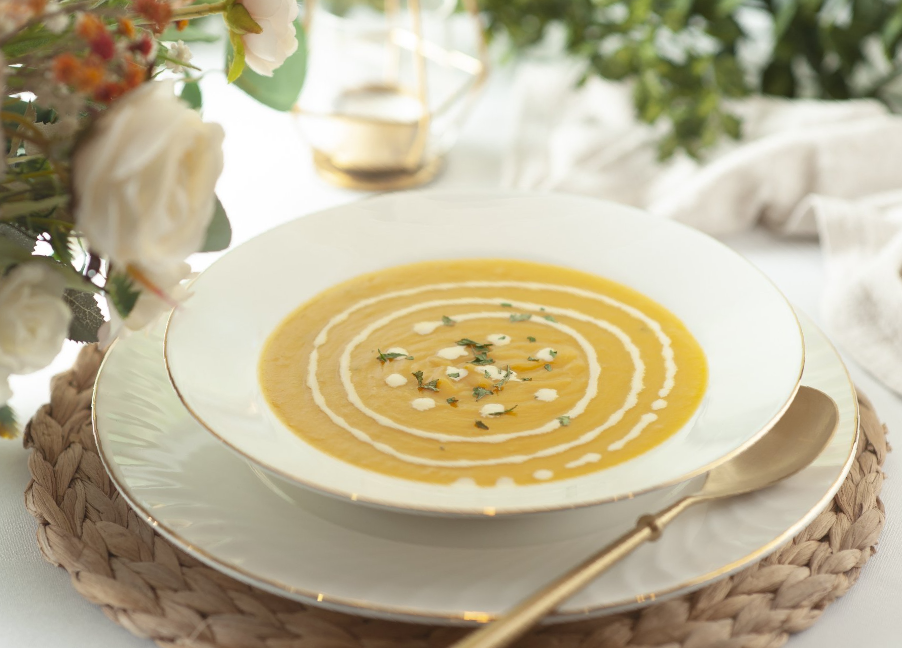 Creamy butternut squash soup in a white bowl, garnished with cream swirls and herbs, on a decorative plate, spoon nearby.