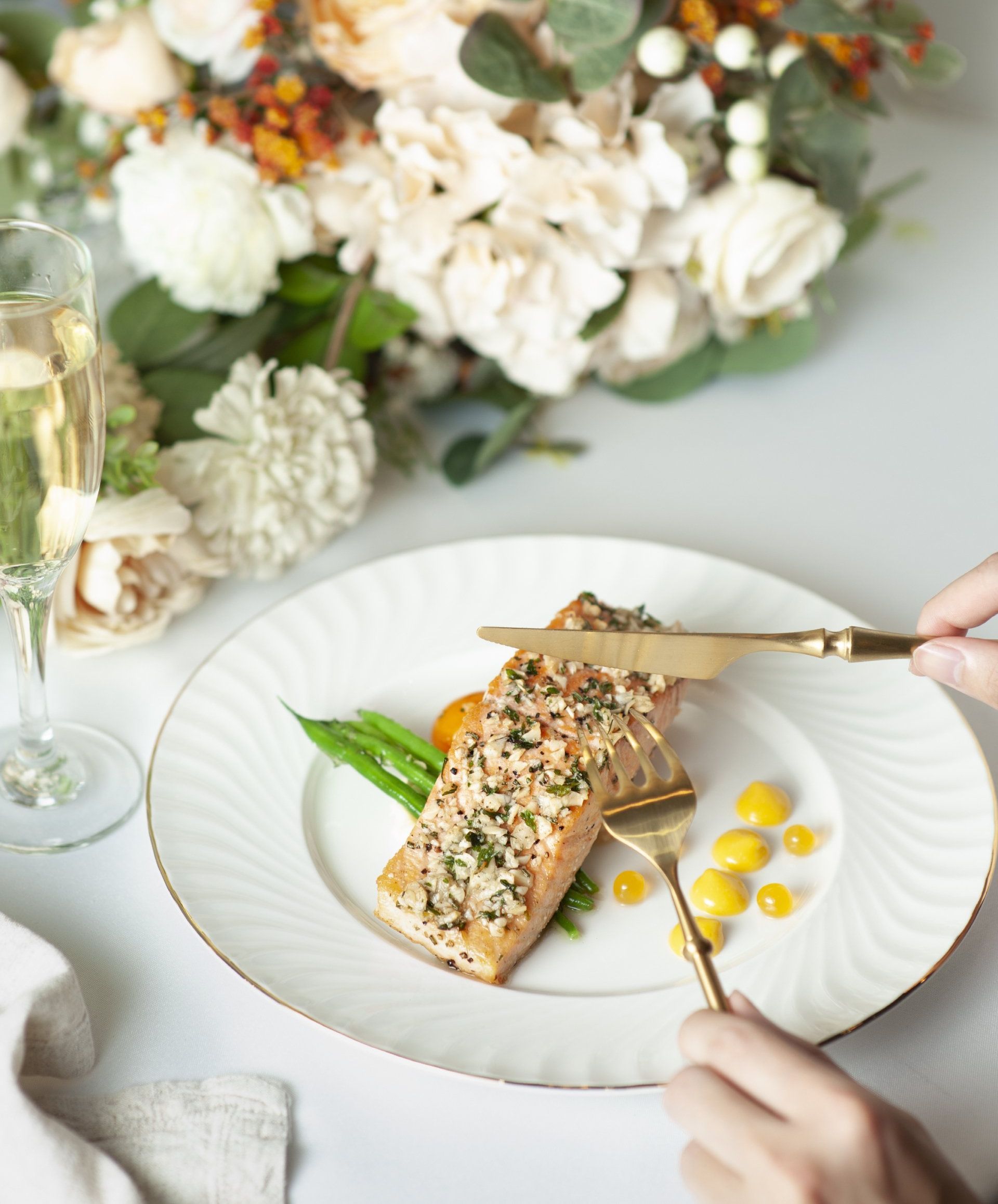 Plate of salmon with asparagus, yellow sauce, and a person using gold silverware near flowers.