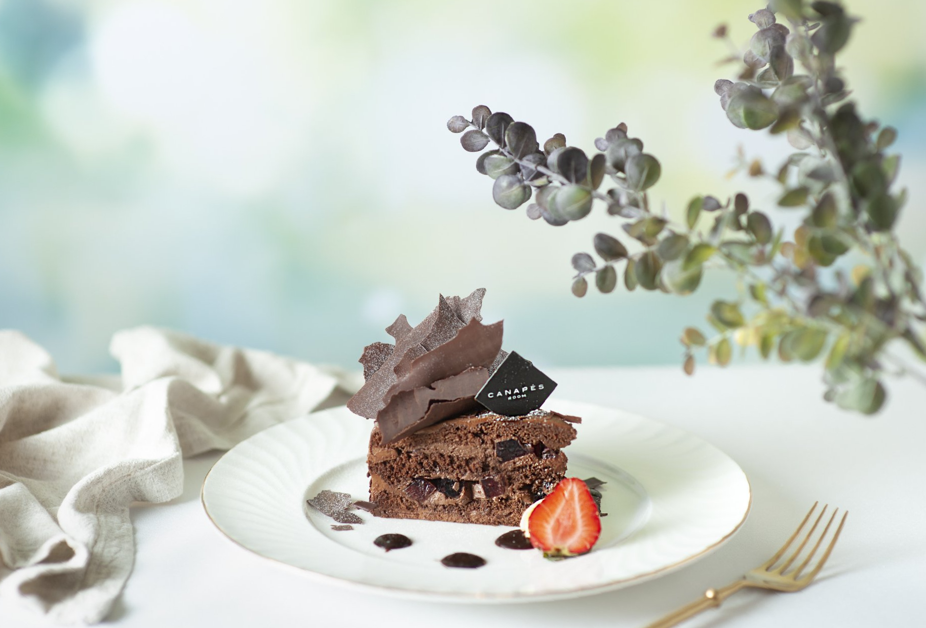 Chocolate cake slice on a white plate, with chocolate shards, a strawberry, and foliage in the background.