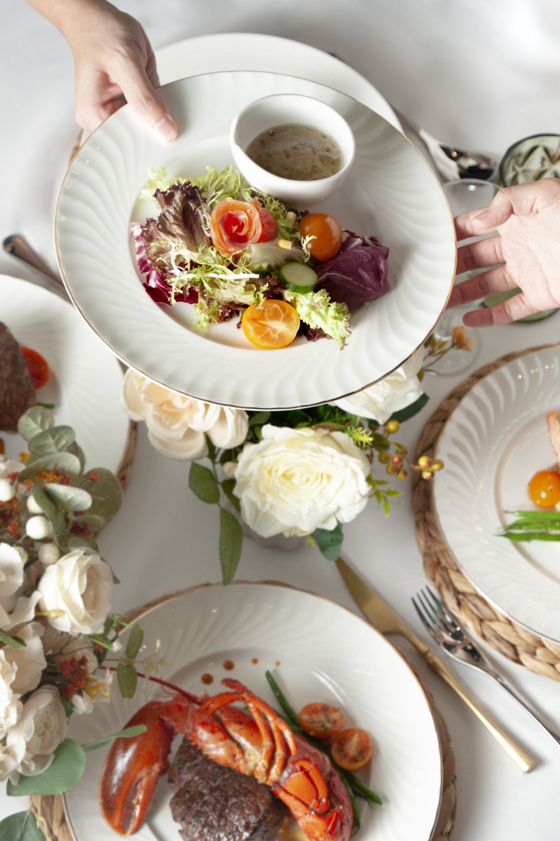 Person holding a plate of salad with dressing; elegant dining table with other dishes.