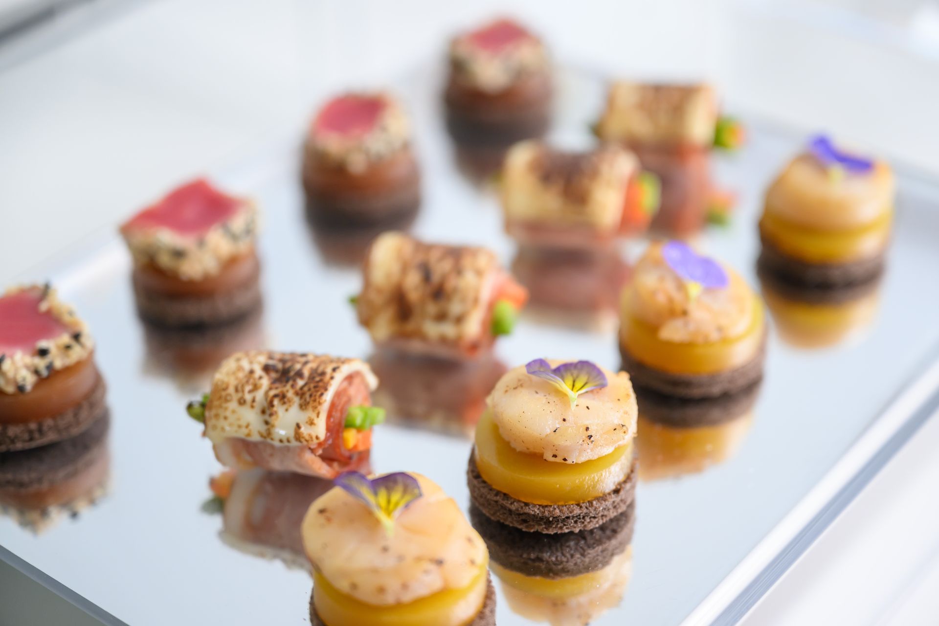 Tray of assorted appetizers, including tuna bites, wraps, and macarons, on a reflective surface.