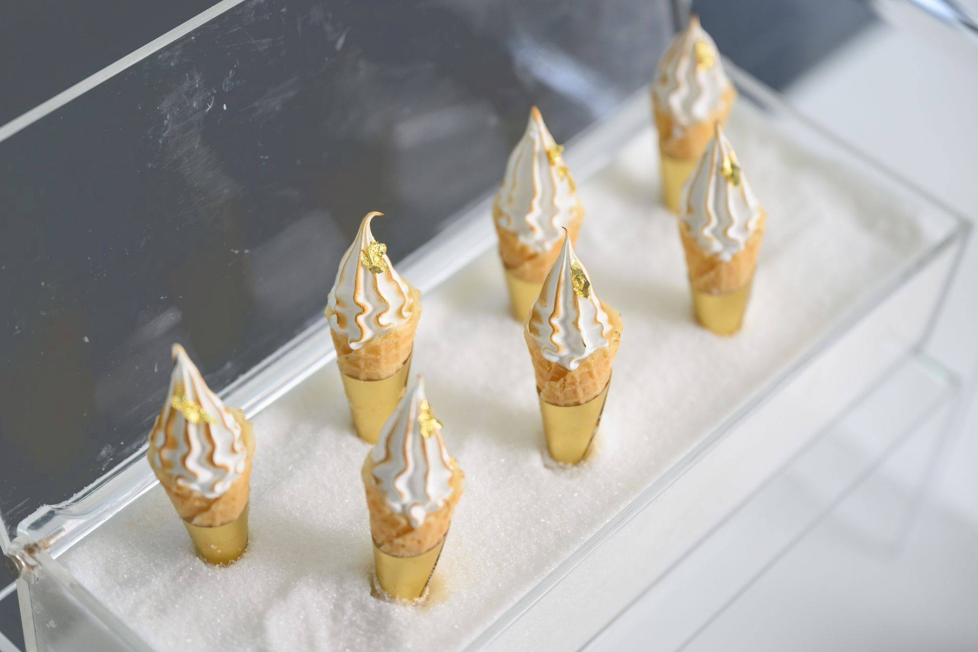 Seven dessert cones with white frosting and gold leaf, arranged on a white tray.
