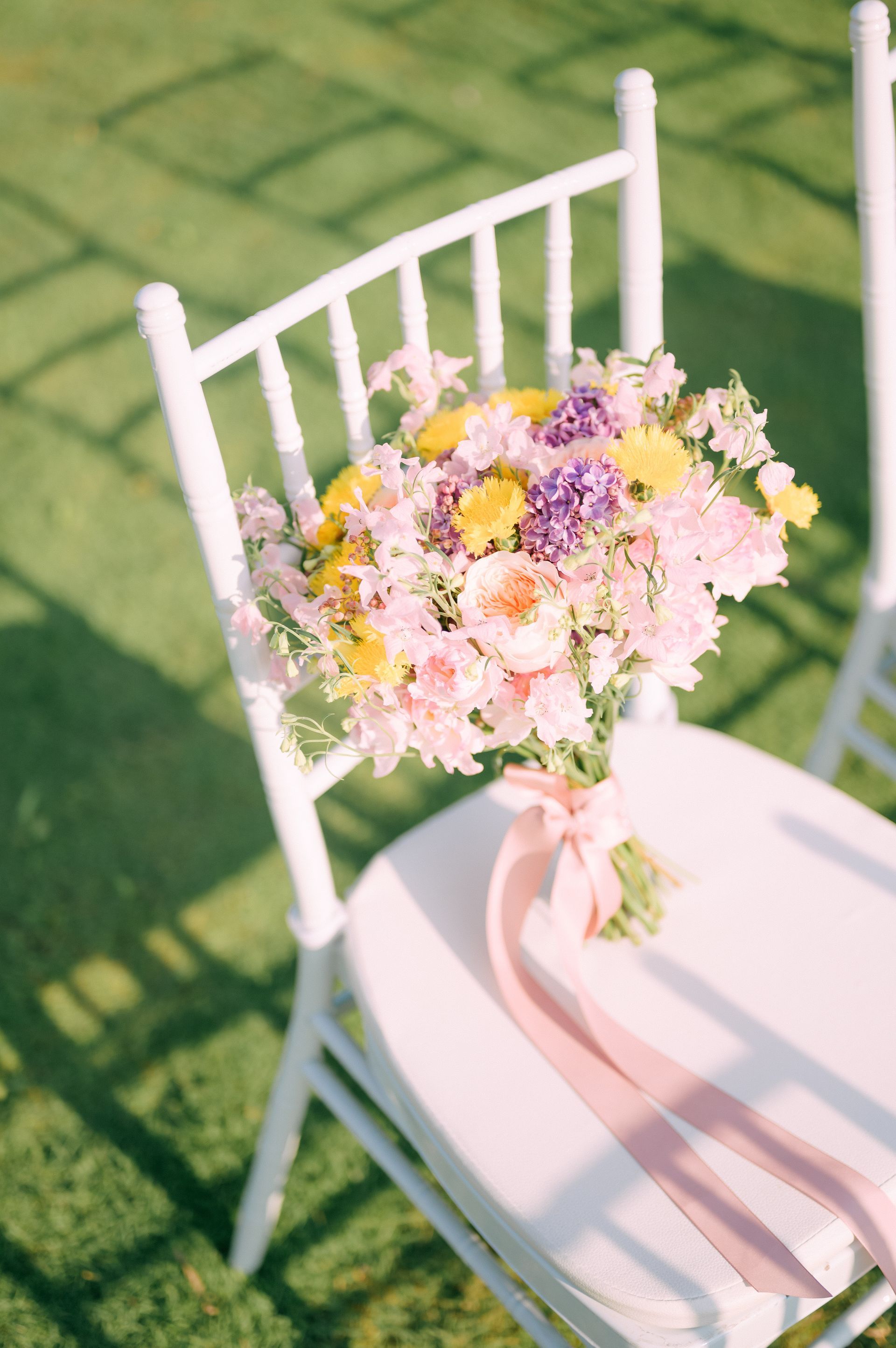 Bouquet of pink, yellow, and purple flowers tied with a pink ribbon, resting on a white chair in a grassy setting.
