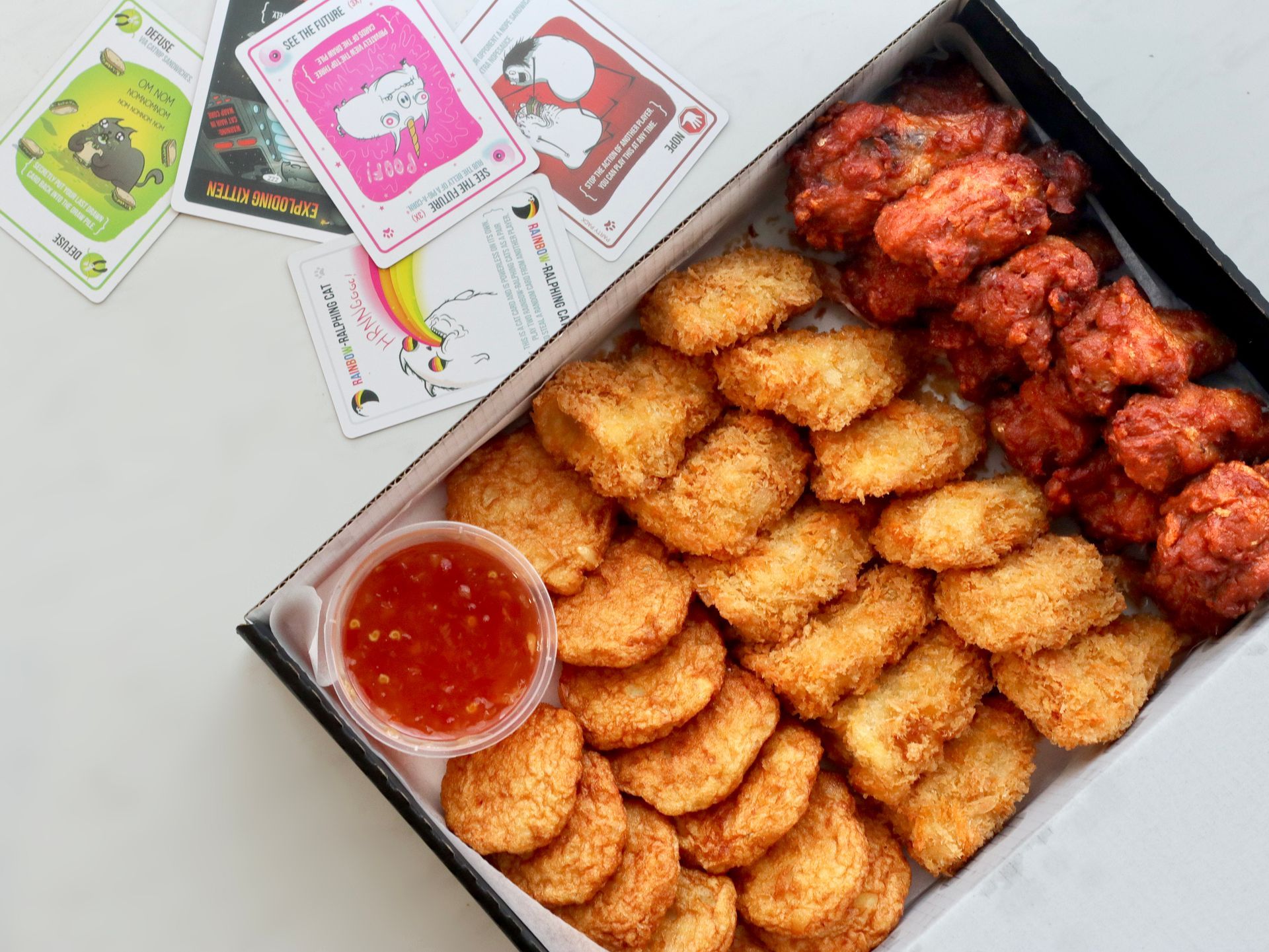 Box of fried chicken nuggets and spicy wings with dipping sauce and card game.