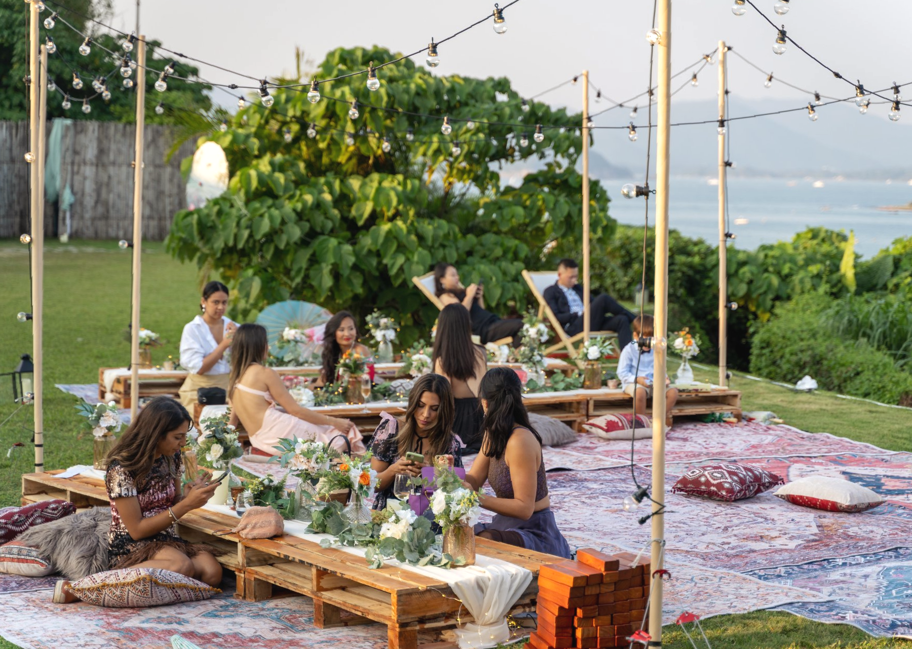 Outdoor picnic with people at low tables, string lights, and a scenic view.