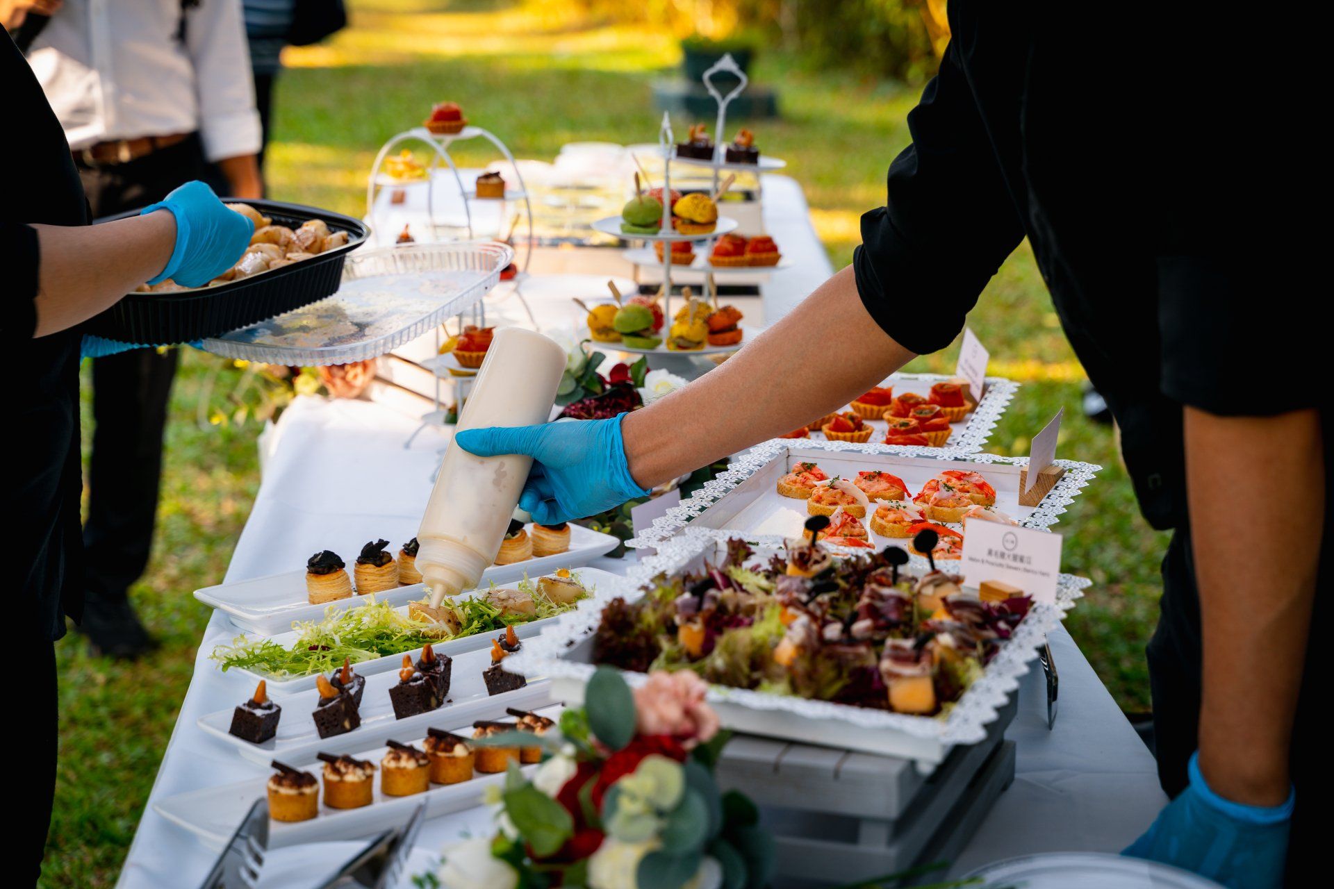 People preparing and serving food on a buffet table outdoors, with white tablecloths and various appetizers.