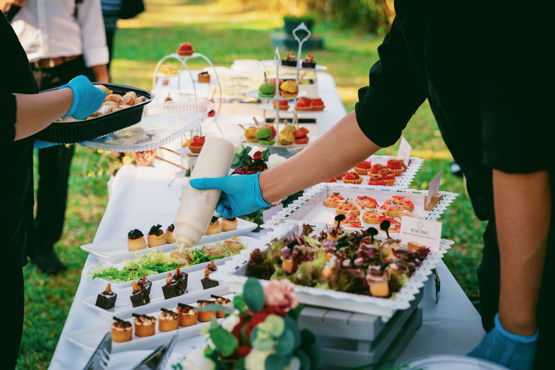 Caterers serving various appetizers on a table outdoors; one squeezes sauce, wearing blue gloves.