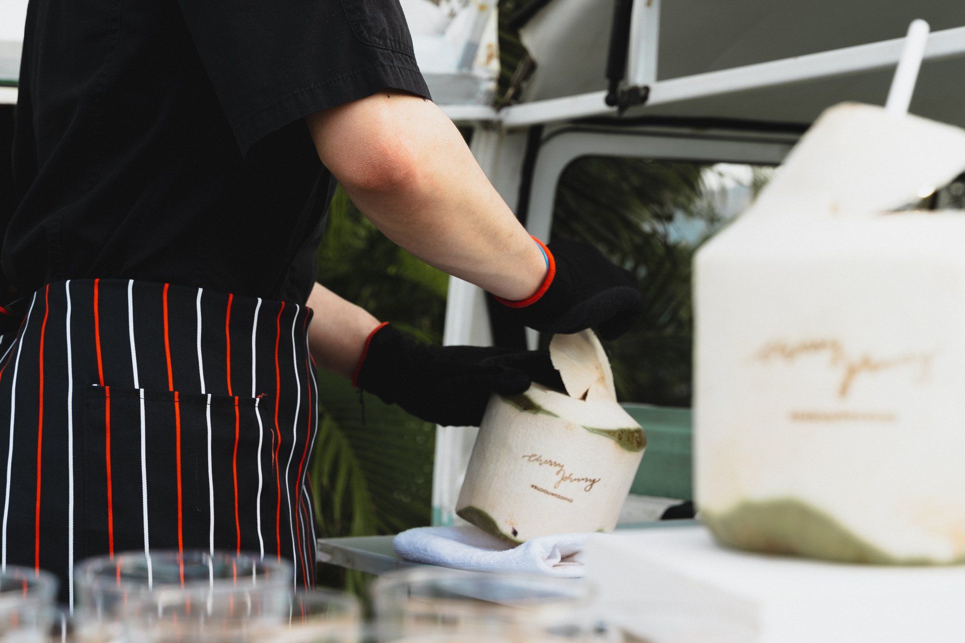 Person in black gloves opening a coconut in front of a food truck.